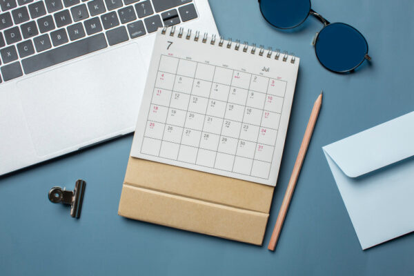 The image presents a neatly arranged workspace featuring a laptop, a paper calendar with dates marked, a pencil, sunglasses, an envelope, and a couple of notebooks on a blue background, symbolizing organization and planning.